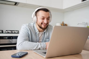 Evde dizüstü bilgisayarı ve kulaklığı olan genç adam, çalışıyor, serbest çalışıyor.