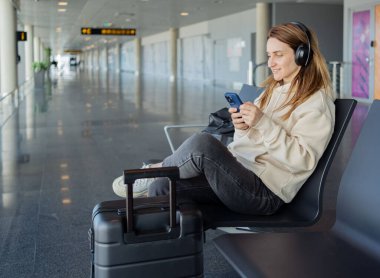 Kadın uçuşunu beklerken akıllı telefon ve kulaklık kullanıyor. Öğrenci kız havaalanında müzik dinliyor ya da podcast yapıyor..