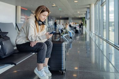 Kadın uçuşunu beklerken akıllı telefon ve kulaklık kullanıyor. Öğrenci kız havaalanında müzik dinliyor ya da podcast yapıyor..