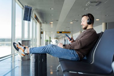 Genç adam havaalanında uçuşu bekliyor. Adam kulaklıktan müzik dinliyor ve akıllı telefon kullanıyor..