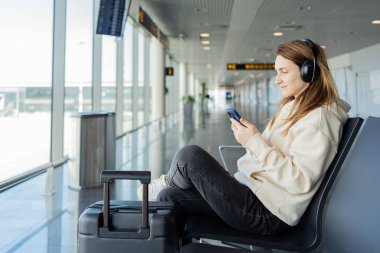 Kadın uçuşunu beklerken akıllı telefon ve kulaklık kullanıyor. Öğrenci kız havaalanında müzik dinliyor ya da podcast yapıyor..