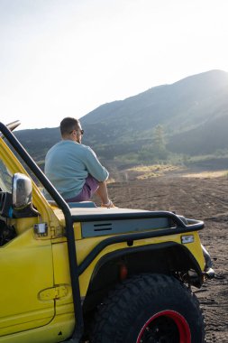 Güzel kayalık manzaraların tadını çıkaran bir adam yol kenarındaki arabanın kaputunda oturuyor. Bali 'deki volkanik vadide seyahat ederken.