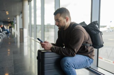 Havalimanında çanta ve cep telefonuyla rahatlayan neşeli bir adamın portresi, sosyal medyada cep telefonuyla iletişim kuran, internetten bilgi arayan, uçuş için bekleyen bir adam.. 