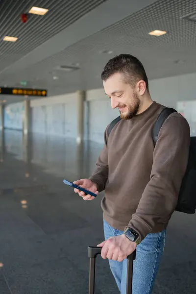 バッグと携帯電話で空港でリラックスする陽気な男の肖像画 男はソーシャルメディアで通信する携帯電話を使用してフライトを待って オンラインで情報を検索 — ストック写真