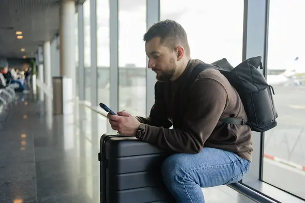 portrait of cheerful man relaxing at airport with bag and mobile phone, guy waiting for flight using mobile phone communicating in social media, searching information online.