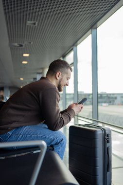 Havalimanında çanta ve cep telefonuyla rahatlayan neşeli bir adamın portresi, sosyal medyada cep telefonuyla iletişim kuran, internetten bilgi arayan, uçuş için bekleyen bir adam.. 