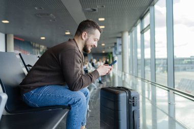 Havalimanında çanta ve cep telefonuyla rahatlayan neşeli bir adamın portresi, sosyal medyada cep telefonuyla iletişim kuran, internetten bilgi arayan, uçuş için bekleyen bir adam.. 
