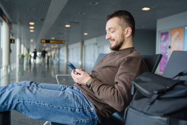 Havalimanında çanta ve cep telefonuyla rahatlayan neşeli bir adamın portresi, sosyal medyada cep telefonuyla iletişim kuran, internetten bilgi arayan, uçuş için bekleyen bir adam.. 