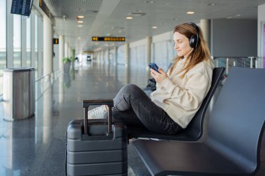 Kadın uçuşunu beklerken akıllı telefon ve kulaklık kullanıyor. Öğrenci kız havaalanında müzik dinliyor ya da podcast yapıyor..