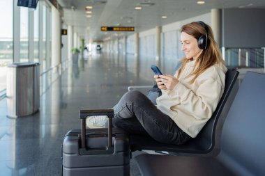 Kadın uçuşunu beklerken akıllı telefon ve kulaklık kullanıyor. Öğrenci kız havaalanında müzik dinliyor ya da podcast yapıyor..