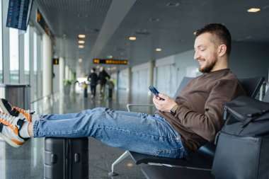 Havalimanında çanta ve cep telefonuyla rahatlayan neşeli bir adamın portresi, sosyal medyada cep telefonuyla iletişim kuran, internetten bilgi arayan, uçuş için bekleyen bir adam.. 