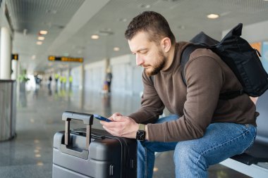Havalimanında çanta ve cep telefonuyla rahatlayan neşeli bir adamın portresi, sosyal medyada cep telefonuyla iletişim kuran, internetten bilgi arayan, uçuş için bekleyen bir adam.. 