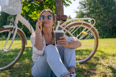 European young woman in park with bicycle and talking on mobile phone. 