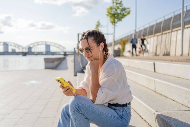 Gülümseyen güzel bir kadın elinde cep telefonuyla müzik dinliyor, sokakta video seyrediyor. Gezici uygulama alışverişini açık havada yapan mutlu hippi kadın..