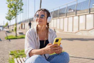 Bluetooth kulaklıklı hipster kadın şehirdeki 4G kablosuz bağlantısını kullanarak mobil radyodan pozitif müzik listesinin keyfini çıkarıyor, video görüşmesi yapıyor, elinde kahve tutan bir kız var ve akıllı telefon kullanıyor..