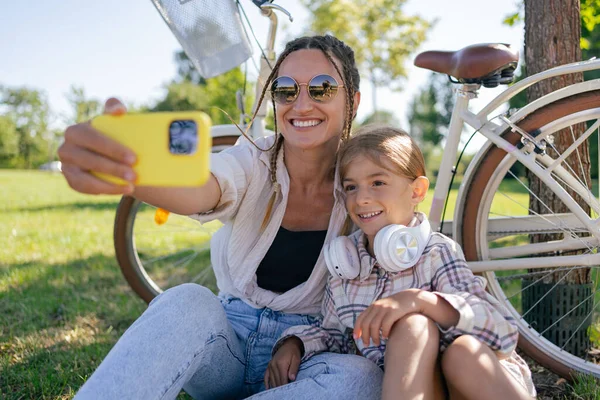 family, leisure and technology concept - happy mother and little daughter with smartphone and bicycles in summer park. Woman and child make selfie in park