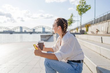 Gülümseyen güzel bir kadın elinde cep telefonuyla müzik dinliyor, sokakta video seyrediyor. Gezici uygulama alışverişini açık havada yapan mutlu hippi kadın..
