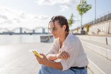 Gülümseyen güzel bir kadın elinde cep telefonuyla müzik dinliyor, sokakta video seyrediyor. Gezici uygulama alışverişini açık havada yapan mutlu hippi kadın..