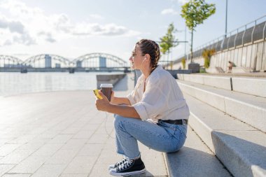 Gülümseyen güzel bir kadın elinde cep telefonuyla müzik dinliyor, sokakta video seyrediyor. Gezici uygulama alışverişini açık havada yapan mutlu hippi kadın..