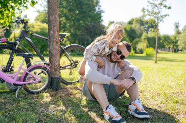 Gülen baba ve kızı yeşil parkta birlikte eğleniyor, rahatlıyor ve bisiklet sürdükten sonra birlikte eğleniyorlar. .