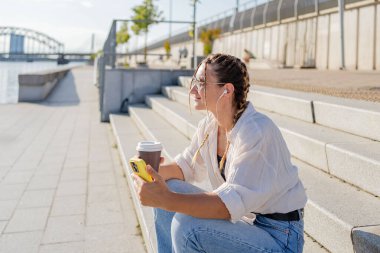 Gülümseyen güzel bir kadın elinde cep telefonuyla müzik dinliyor, sokakta video seyrediyor. Gezici uygulama alışverişini açık havada yapan mutlu hippi kadın..
