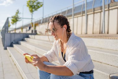 Gülümseyen güzel bir kadın elinde cep telefonuyla müzik dinliyor, sokakta video seyrediyor. Gezici uygulama alışverişini açık havada yapan mutlu hippi kadın..