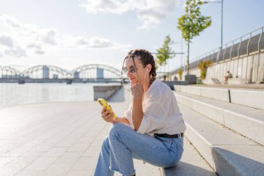 Gülümseyen güzel bir kadın elinde cep telefonuyla müzik dinliyor, sokakta video seyrediyor. Gezici uygulama alışverişini açık havada yapan mutlu hippi kadın..