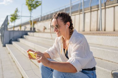 Gülümseyen güzel bir kadın elinde cep telefonuyla müzik dinliyor, sokakta video seyrediyor. Gezici uygulama alışverişini açık havada yapan mutlu hippi kadın..