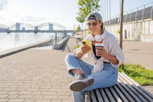 若いヒスパニックヒップスター少女はテイクアウト飲料で通りに座っている間携帯電話をブラウズ — ストック写真