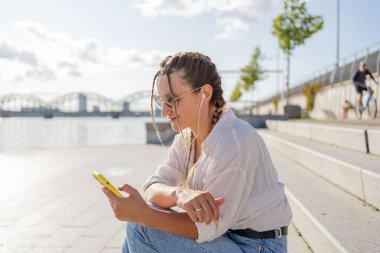Gülümseyen güzel bir kadın elinde cep telefonuyla müzik dinliyor, sokakta video seyrediyor. Gezici uygulama alışverişini açık havada yapan mutlu hippi kadın..