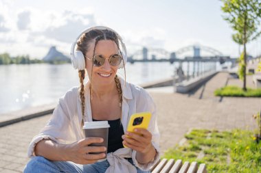 Bluetooth kulaklıklı hipster kadın şehirdeki 4G kablosuz bağlantısını kullanarak mobil radyodan pozitif müzik listesinin keyfini çıkarıyor, video görüşmesi yapıyor, elinde kahve tutan bir kız var ve akıllı telefon kullanıyor..