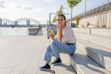 Gülümseyen güzel bir kadın elinde cep telefonuyla müzik dinliyor, sokakta video seyrediyor. Gezici uygulama alışverişini açık havada yapan mutlu hippi kadın..