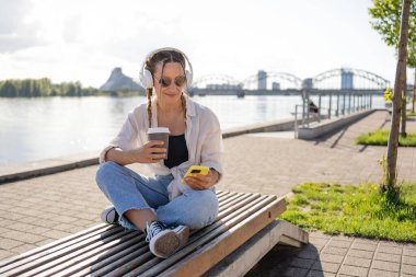 Bluetooth kulaklıklı hipster kadın şehirdeki 4G kablosuz bağlantısını kullanarak mobil radyodan pozitif müzik listesinin keyfini çıkarıyor, video görüşmesi yapıyor, elinde kahve tutan bir kız var ve akıllı telefon kullanıyor..