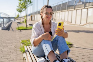 Kuaförlü hippi kadın ve kahve servisi şehrin caddesinde akıllı telefon kullanarak oturuyor. Dışarıda elinde akıllı telefonuyla oturan mutlu öğrenci kız kahve molası veriyor arkadaşlarıyla görüntülü görüşme yapıyor..