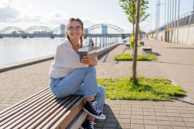 Genç hippi kadın şehirde bir bankta otururken elinde bir fincan kahveyle oturuyor. Güneşli bahar günü