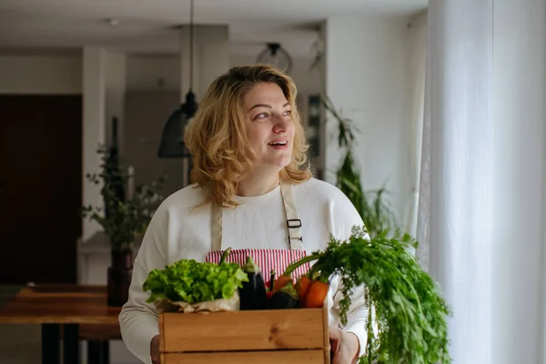 Bir ev hanımı pencereden dışarı bakar ve elinde ahşap bir kutu bitki ve sebze tutar.