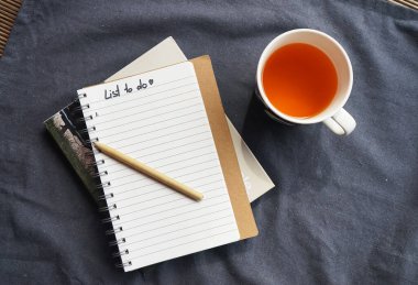 Gri masa örtüsü üzerinde bir bardak çay ve bir kalemle defter.