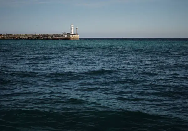 Güneşli bir günde, sakin bir mavi denizin arka planında, taştan bir iskelede beyaz bir deniz feneri duruyor..