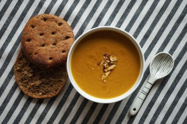 Beyaz tabakta sebze püresi çorbası ve çizgili masa örtüsünde iki dilim ekmek.
