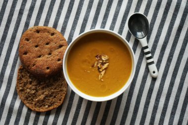 Beyaz tabakta sebze püresi çorbası ve çizgili masa örtüsünde iki dilim ekmek.