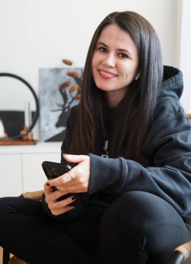 Siyah kapüşonlu siyah saçlı bir kız gülümsüyor ve elinde bir telefon tutuyor.