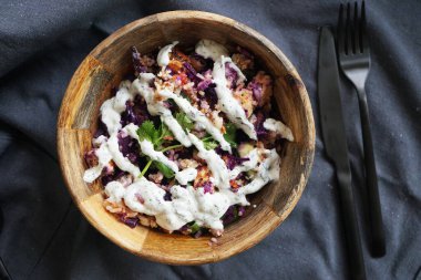 Koyu gri masa örtüsü üzerinde çatal ve bıçakla ahşap bir tabağa sarılı salata. 