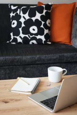 İş yerinde bir dizüstü bilgisayar, bir fincan kahve ve arka planda gri bir kanepe ve çok renkli yastıklar.
