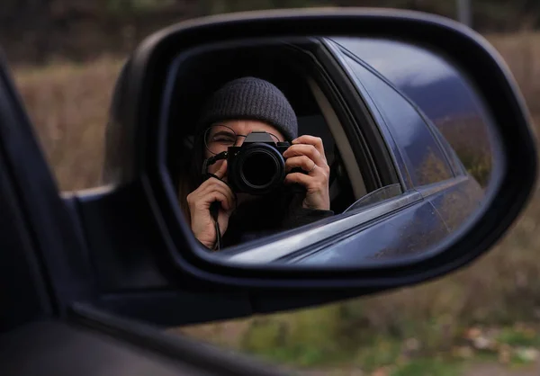 在一个阴郁的秋日里 一个戴着眼镜的黑头发女孩看着摄像机的取景器 并在车镜中反射出自己的影子 — 图库照片