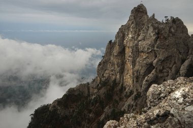 Bulutların üzerinde deniz ve gri gökyüzünün arka planına karşı yüksek bir dağ yükselir.