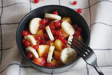 Muzlu, şeftalili ve kırmızı üzümlü meyve salatası siyah bir kasede kareli masa örtüsünde.