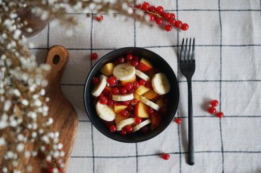 Muzlu, şeftalili ve kırmızı üzümlü meyve salatası siyah bir kasede kareli masa örtüsünde.
