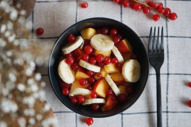 Muzlu, şeftalili ve kırmızı üzümlü meyve salatası siyah bir kasede kareli masa örtüsünde.