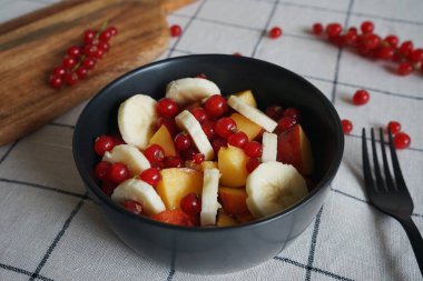 Muzlu, şeftalili ve kırmızı üzümlü meyve salatası siyah bir kasede kareli masa örtüsünde.