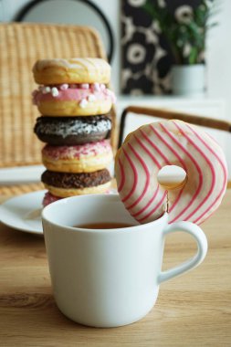 Bir sürü donut pembe ve çikolata kremalı, çok renkli serpiştirilmiş tabakta bir fincan kahvenin yanında bir sandalyenin arka planında yatıyor. 